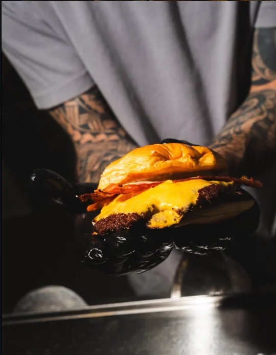Bacon cheeseburger con cheddar fuso tenuto tra due mani con i guanti neri, davanti alla piastra